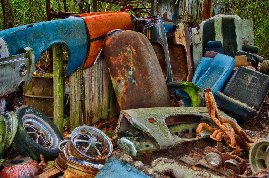 Fotografia przedstawia kolorowe i zardzewiałe elementy karoserii samochodowych, czy też odpadów samochodowych. Jest to zobrazowanie bloga o tytule: "Na czym polega recykling samochodów?".