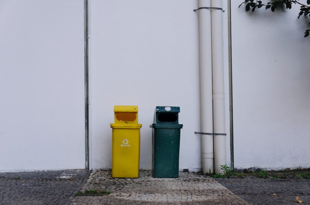 Zdjęcie przedstawia dwa kosze na śmieci. Jeden z nich jest żółty - na plastiki, metale, a drugi czarny - na odpady zmieszane. Fotografia ma przedstawiać problem odpadów zmieszanych - co wrzucać a czego nie?