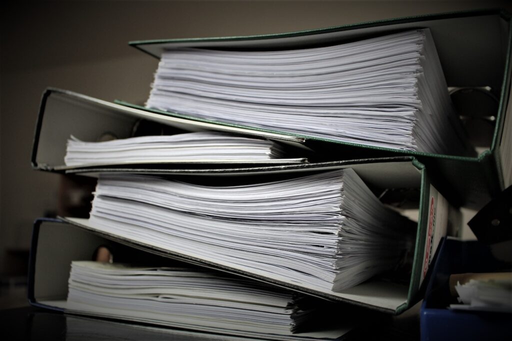 Zdjęcie przedstawia stos segregatorów wypełnionych papierowymi dokumentami. Jest to obrazek dopasowany do bloga o tematyce okresów przechowywania dokumentów - zasad i terminów.