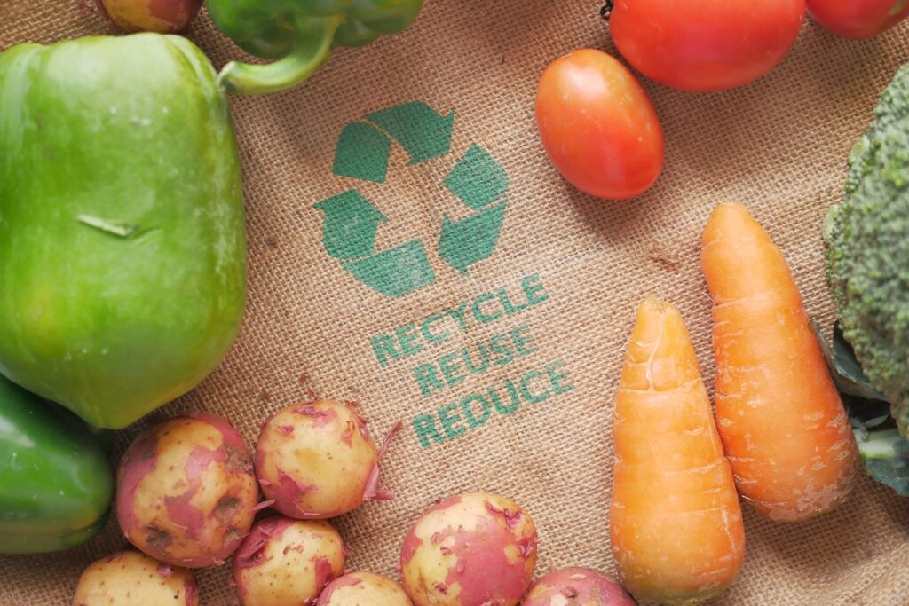 Zdjęcie przedstawia warzywa na jutowej torbie z napisem "Recycle, Reuse, Reduce" i ma stanowić ilustracje do bloga o temacie: "Symbole na opakowaniach i ich znaczenie".