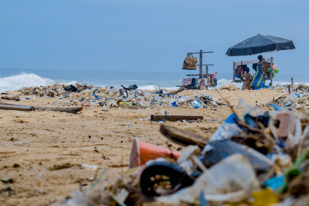Obrazek przedstawia zdjęcie plaży zanieczyszczonej plastikami i różnymi odpadami. Cała fotografia ma obrazować bloga o tytule: "Recykling butelek PET. Na czym polega?".