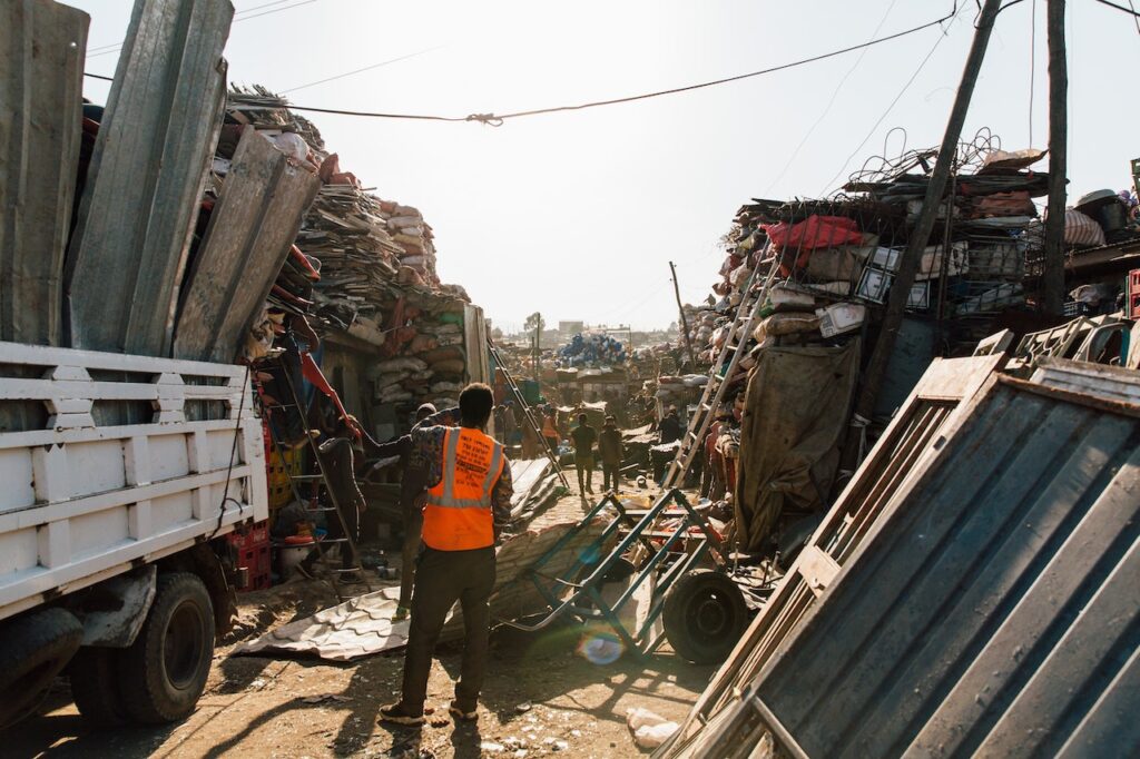 Fotografia przedstawia robotników na złomowisku, w rogu kadru z lewej znajduje się ciężarówka załadowana odpadami. Jest to ilustracja do tekstu o tytule: "Jak wygląda transport złomu?".